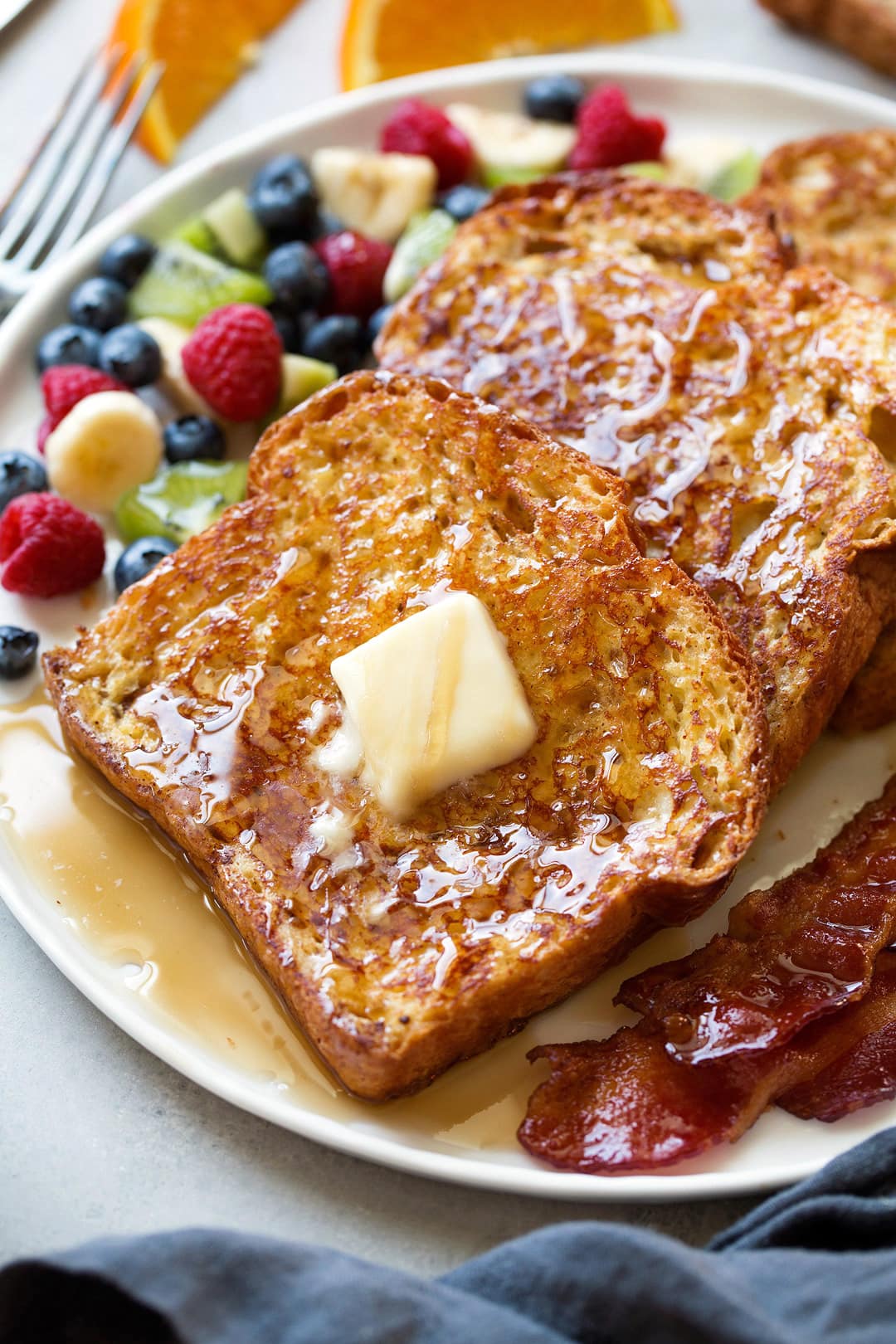 Row of french toast on a plate topped with maple syrup and butter and served with a side of bacon.