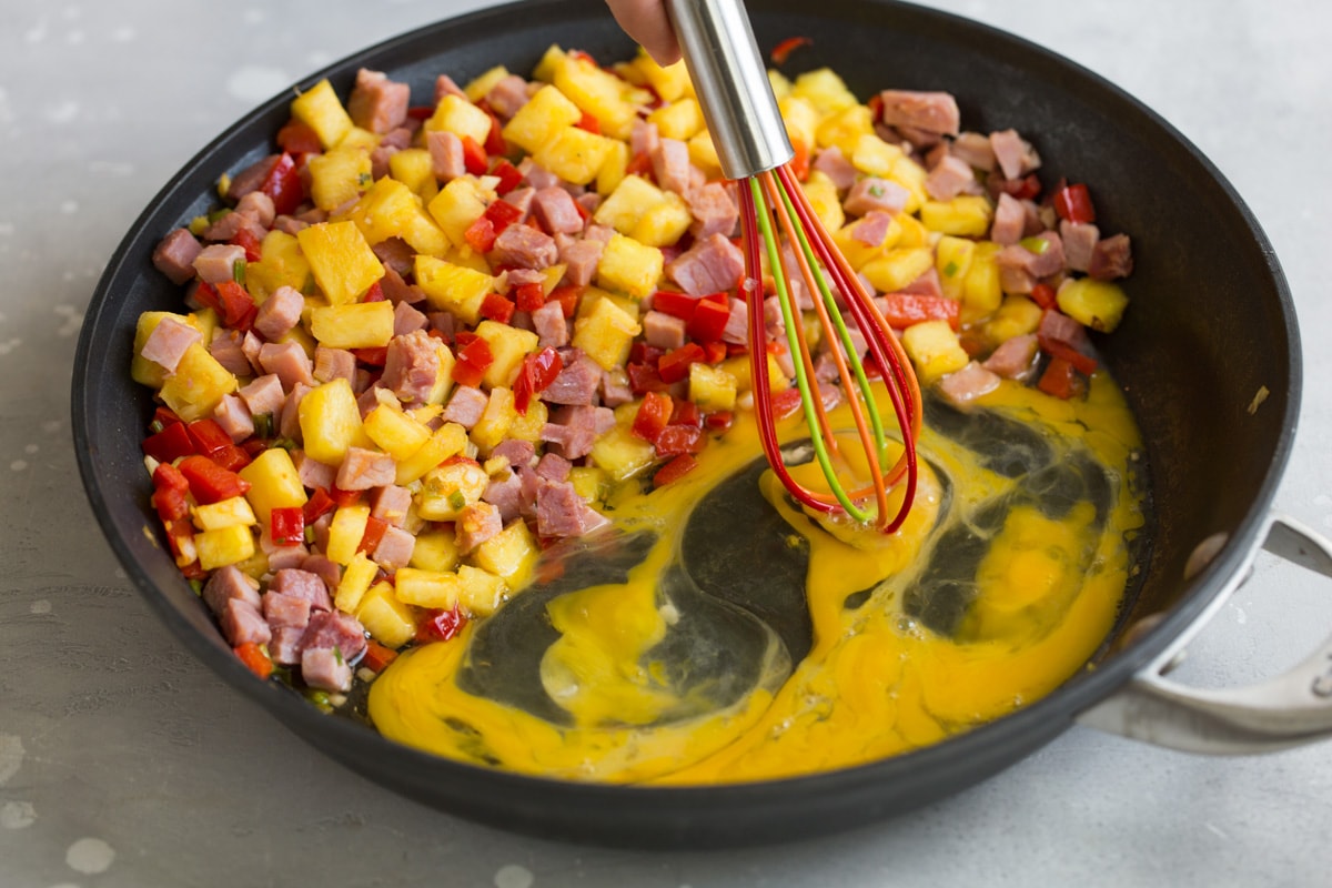 whisking eggs in a skillet next to pineapple ham and bell pepper mixture