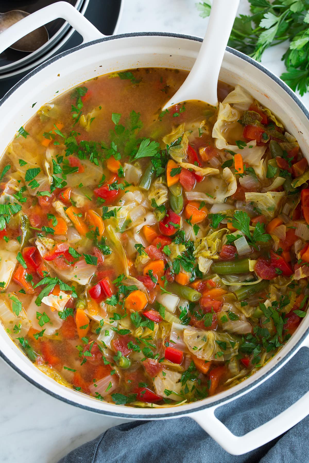 Overhead image of cabbage soup in a large white pot with a white ladle, showing an abundance of fresh vegetables.