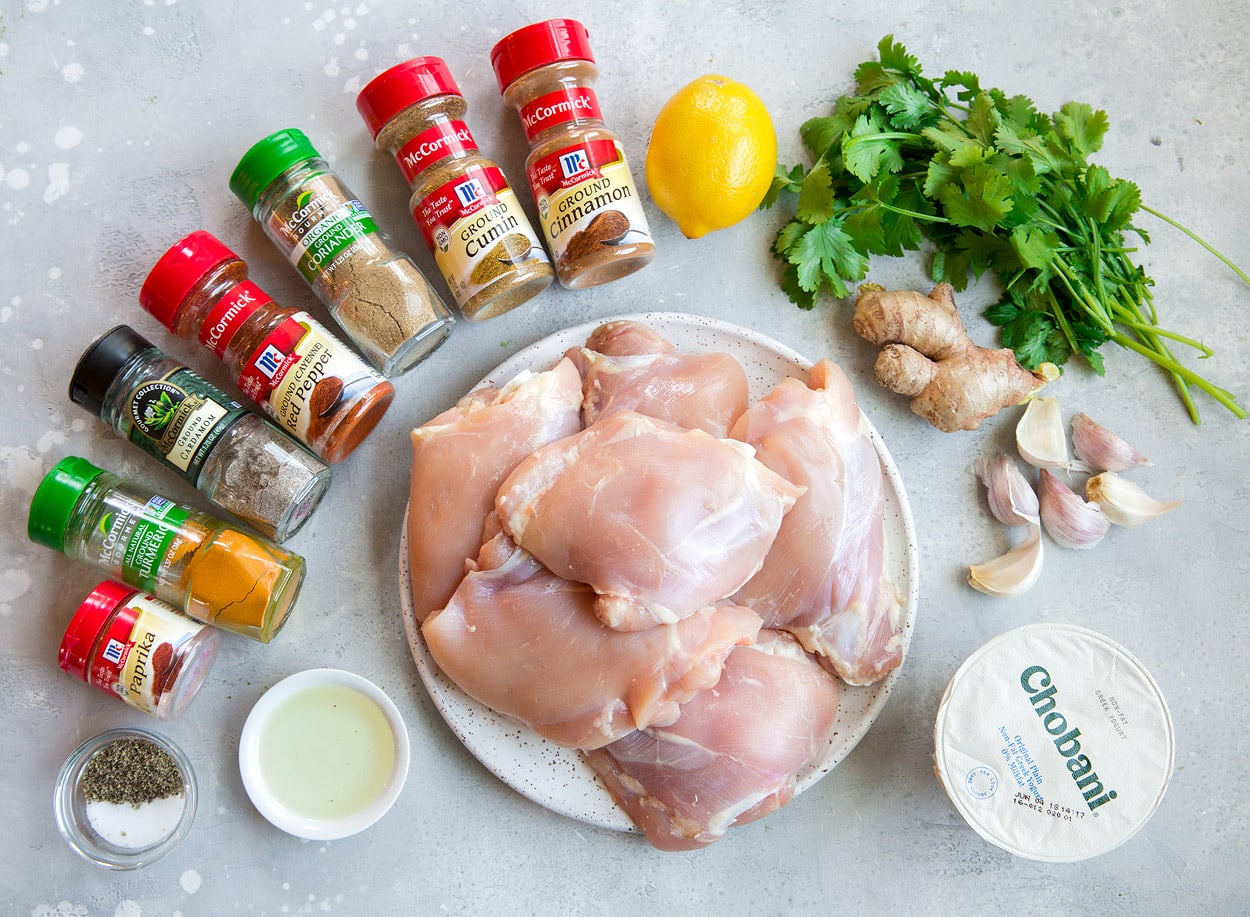 Tandoori Chicken Ingredients including spices, chicken thighs, oil, lemon, cilantro, garlic and greek yogurt shown here on a grey surface.