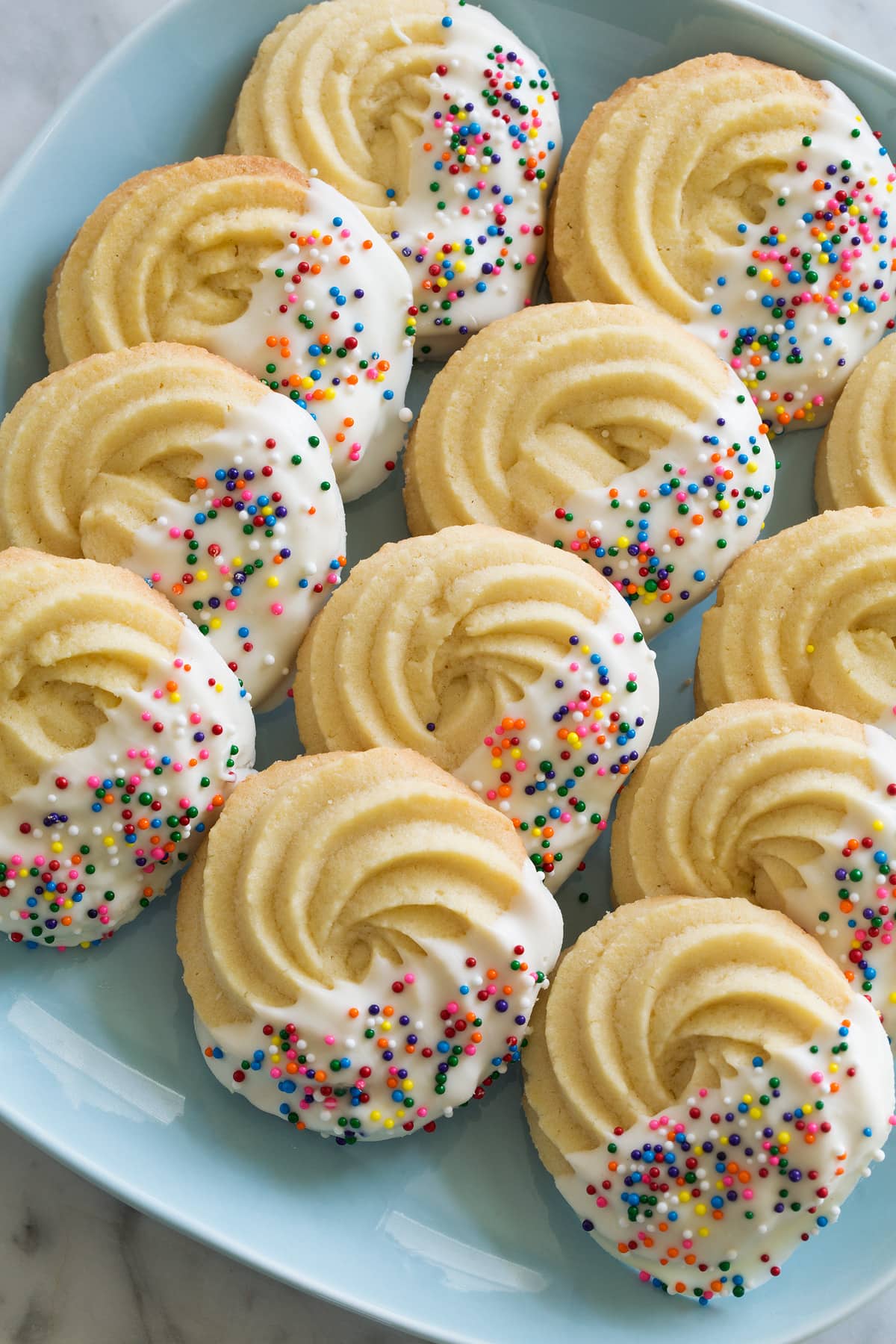 Butter Cookies dipped in white chocolate and covered with rainbow sprinkles. Set in three rows on a turquoise plate. 
