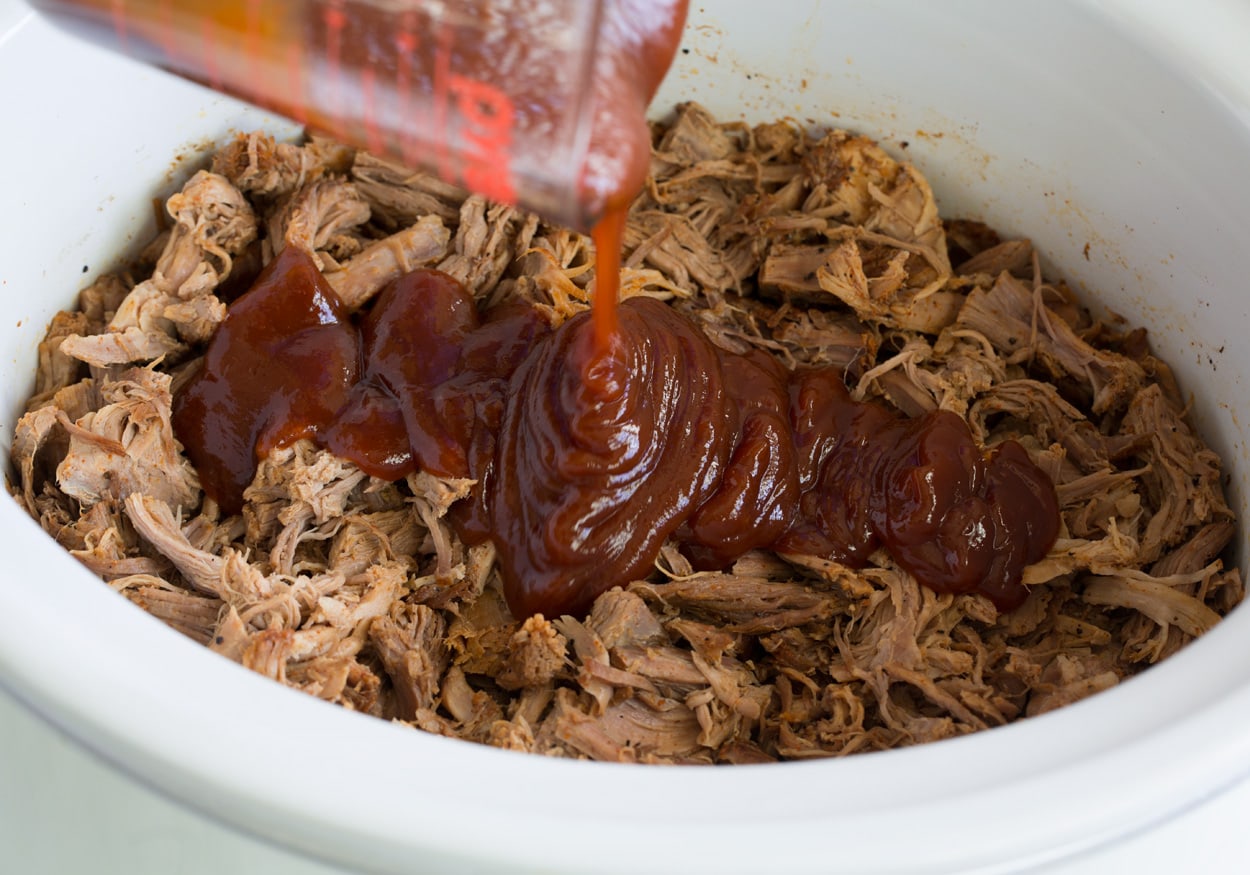 Pulled pork shown here in crock pot pouring barbecue sauce over top.