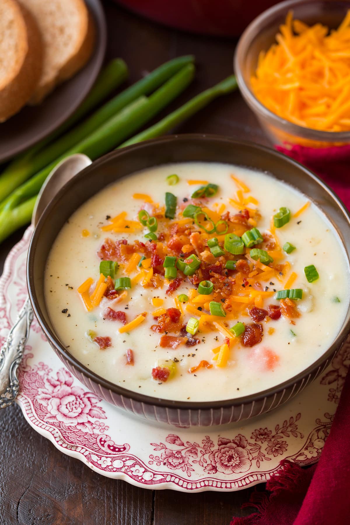 Photo: Serving of potato soup shown in a brown bowl set over a decorated white and red plate. Potato soup is garnished with cheddar, bacon and green onions. Wheat bread is shown as a serving suggestion.