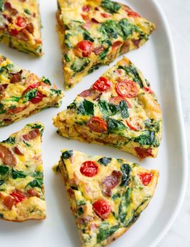 Six frittata wedges shown on a white serving platter set over a marble surface. Frittata is filled with bacon, spinach, tomatoes and swiss.