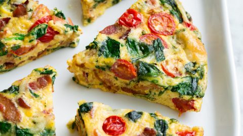 Six frittata wedges shown on a white serving platter set over a marble surface. Frittata is filled with bacon, spinach, tomatoes and swiss.