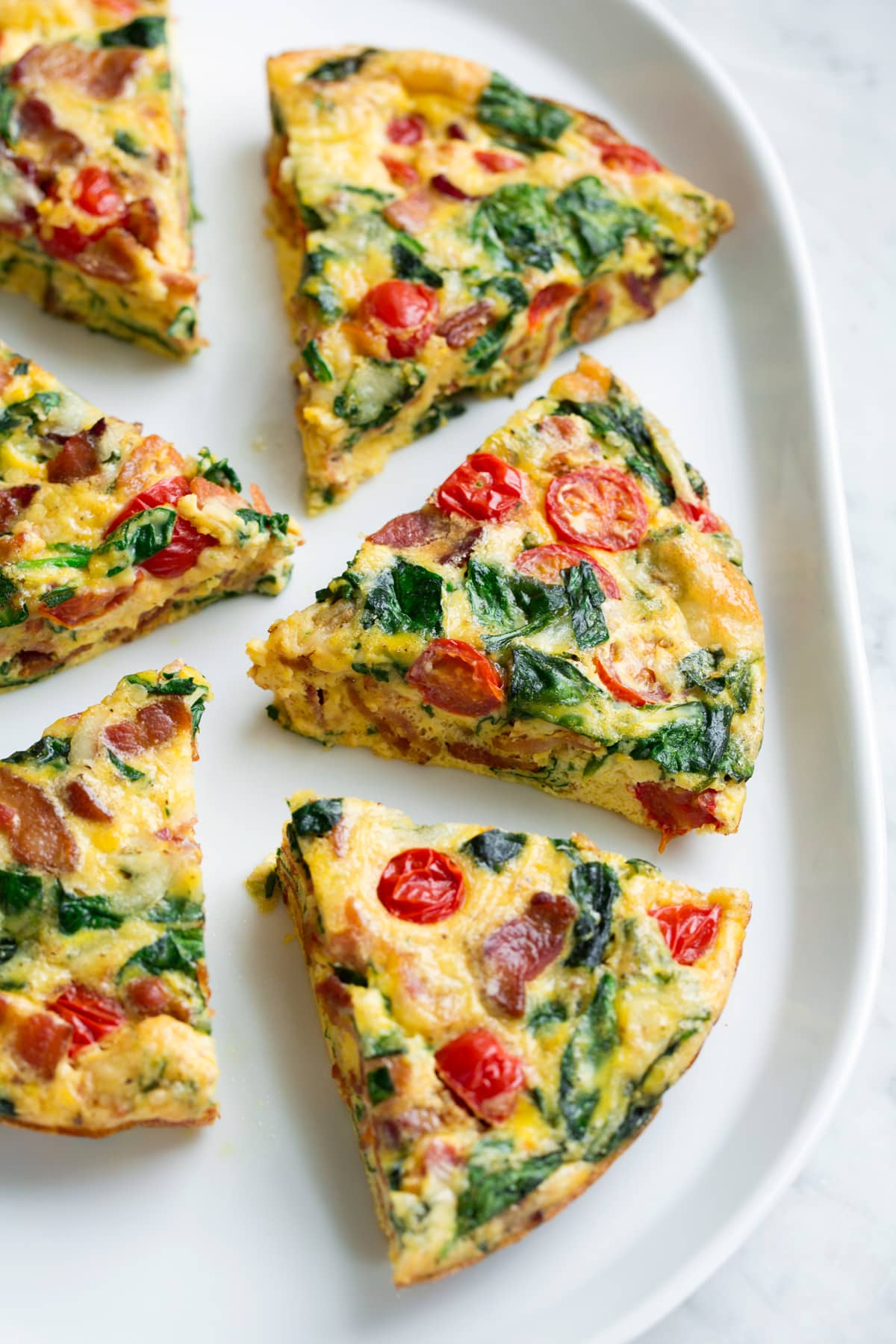 Six frittata wedges shown on a white serving platter set over a marble surface. Frittata is filled with bacon, spinach, tomatoes and swiss.