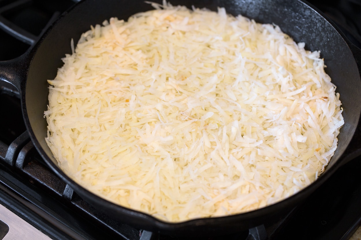 Cooking shredded potatoes in a cast iron skillet over a gas stove top to make hash browns.