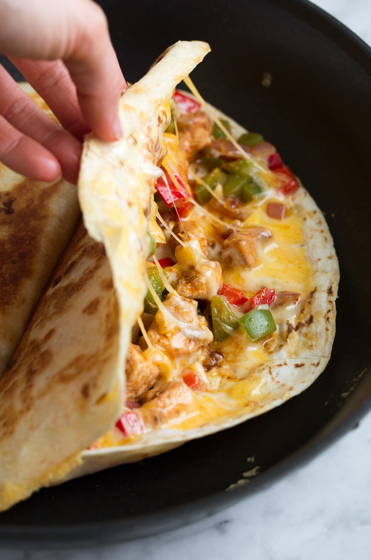 Hand opening quesadilla in a skillet to show filling of cheese, bell peppers, onions, and seasoned chicken pieces.