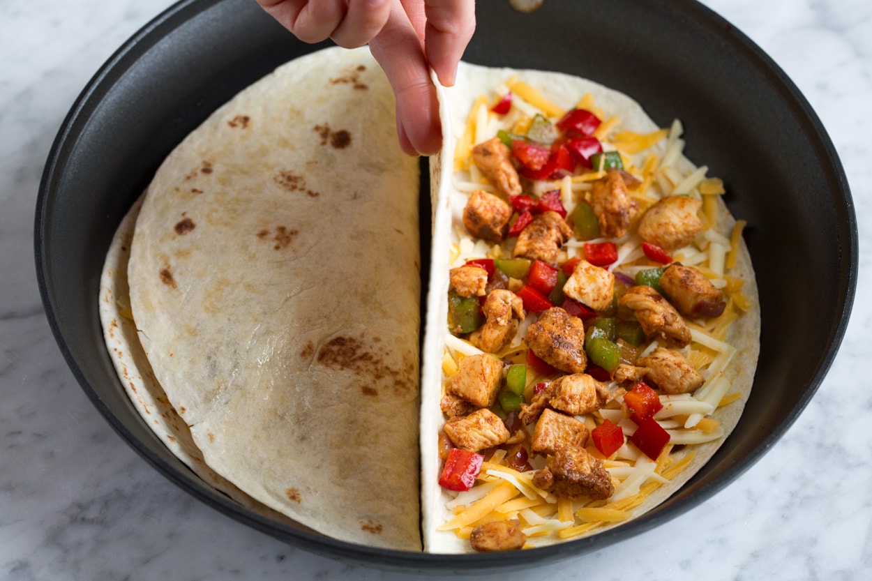Showing how to cook quesadillas, two at a time in a skillet. Layering tortilla then filling half with cheese and fillings and folding over.