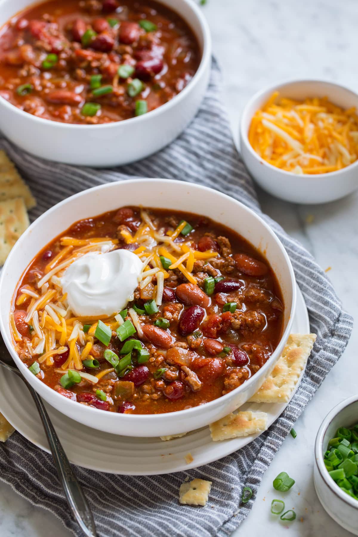 Two servings of chili in white bowls set over a marble surface. Servings topped with cheddar, sour cream and green onions.