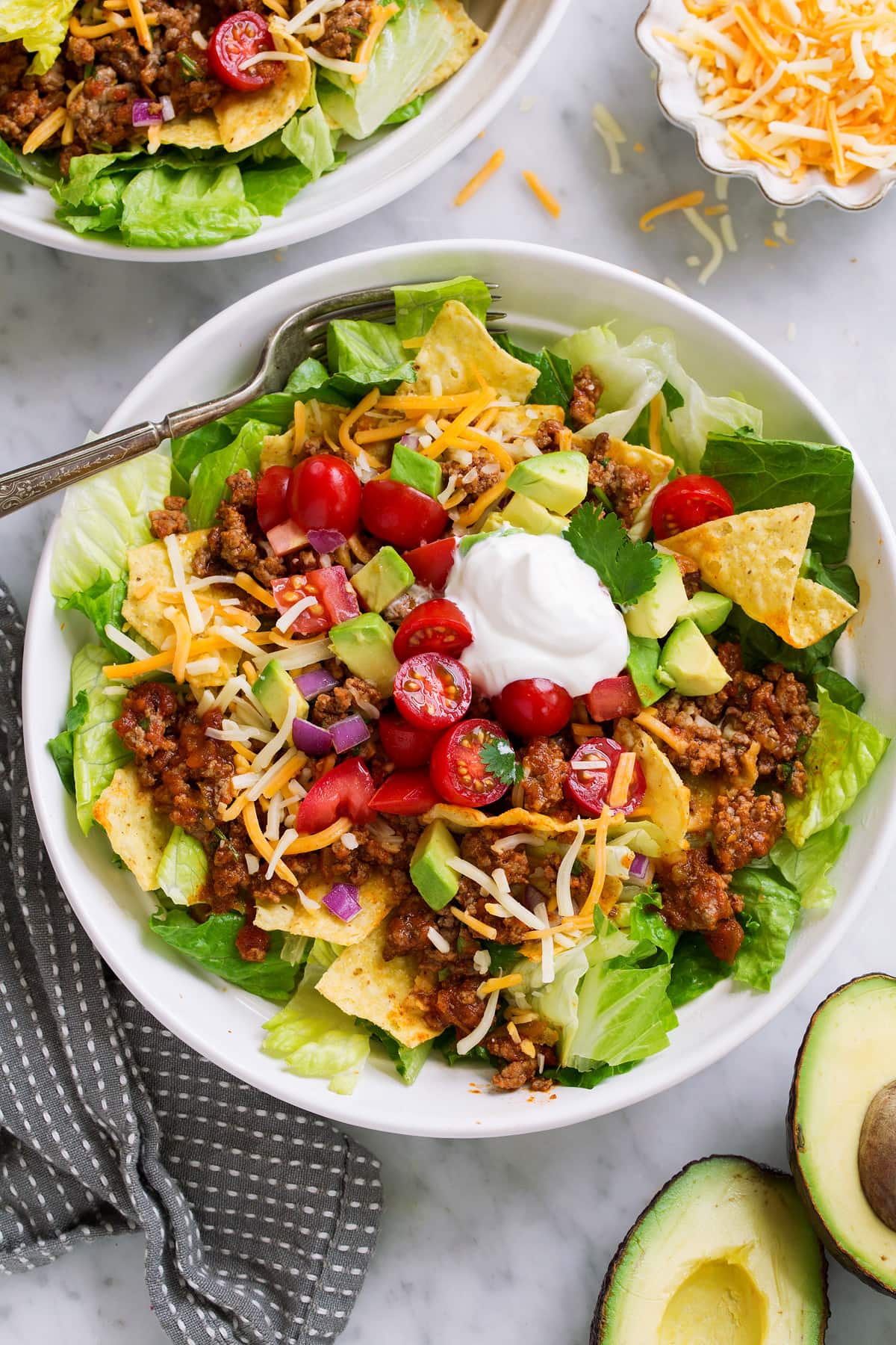 Taco Salad in a white individual serving bowl set over a marble surface. Taco salad is layered with romaine lettuce, seasoned ground beef, cheese, tomatoes, avocado and sour cream.
