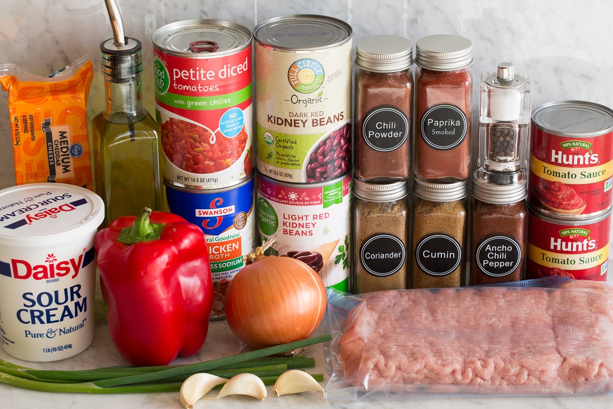 Photo of ingredients used to make turkey chili. Includes ground turkey, tomato sauce, chili powder, ancho chili powder, smoked paprika, cumin, coriander, light and dark red kidney beans, chicken broth, canned tomatoes, bell pepper, yellow onion, green onions, garlic, olive oil, sour cream, and cheddar cheese.