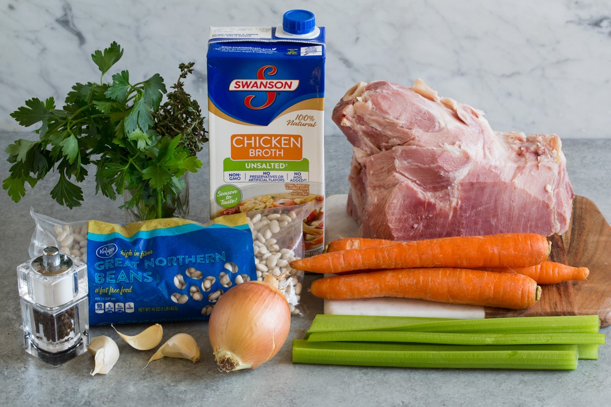 Ingredients needed for ham and bean soup shown here including meaty ham bone, carrots, celery, onion, garlic, great northern beans, chicken broth, parsley and thyme.