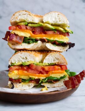 Bagel sandwich with microwave eggs, bacon, avocado, tomato, cheddar and lettuce. Sandwich is cut in half and stacked on a wooden plate set over a marble surface.