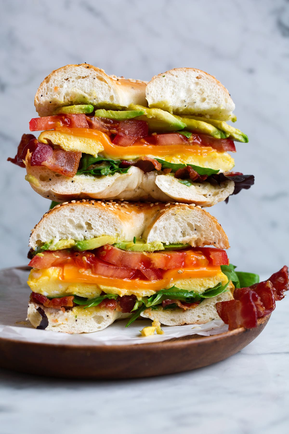 Bagel sandwich with microwave eggs, bacon, avocado, tomato, cheddar and lettuce. Sandwich is cut in half and stacked on a wooden plate set over a marble surface.