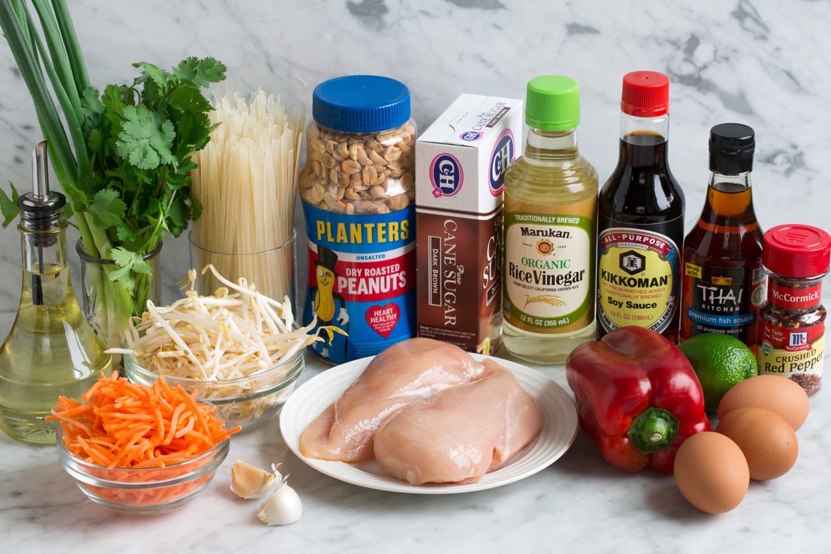 Ingredients needed for pad thai shown here including eggs, red pepper flakes, fish sauce, soy sauce, rice vinegar, lime, bell pepper, chicken, brown sugar, peanuts, rice noodles, cilantro, green onions, bean sprouts, carrots, garlic and vegetable oil.