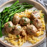 Serving of Swedish meatballs on egg noodles with a side of asparagus on a speckled cream plate.