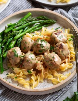 Serving of Swedish meatballs on egg noodles with a side of asparagus on a speckled cream plate.
