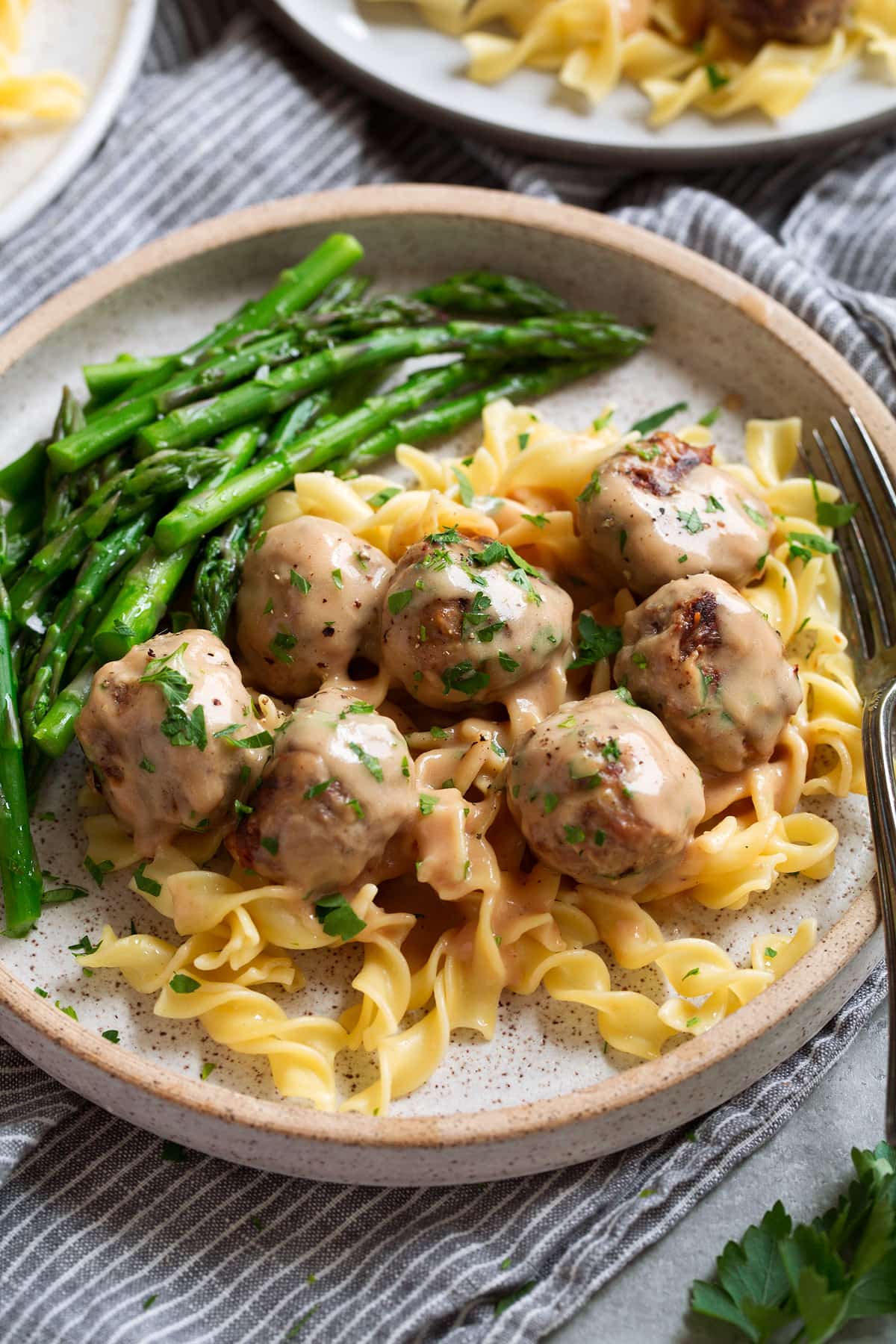 Serving of Swedish meatballs in egg noodles with a side of asparagus on a plate of speckled cream.