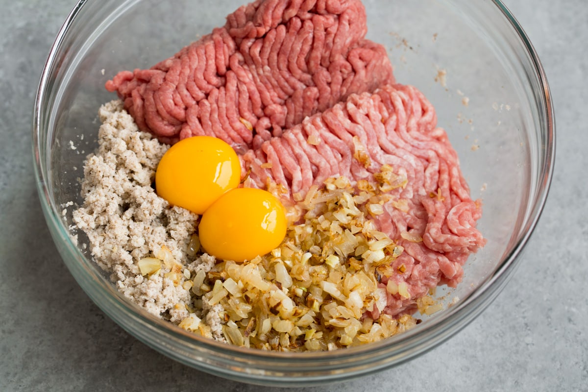 Mixing ground beef, ground pork, bread crumb mixture, sautéed onions and egg yolks in a glass mixing bowl.