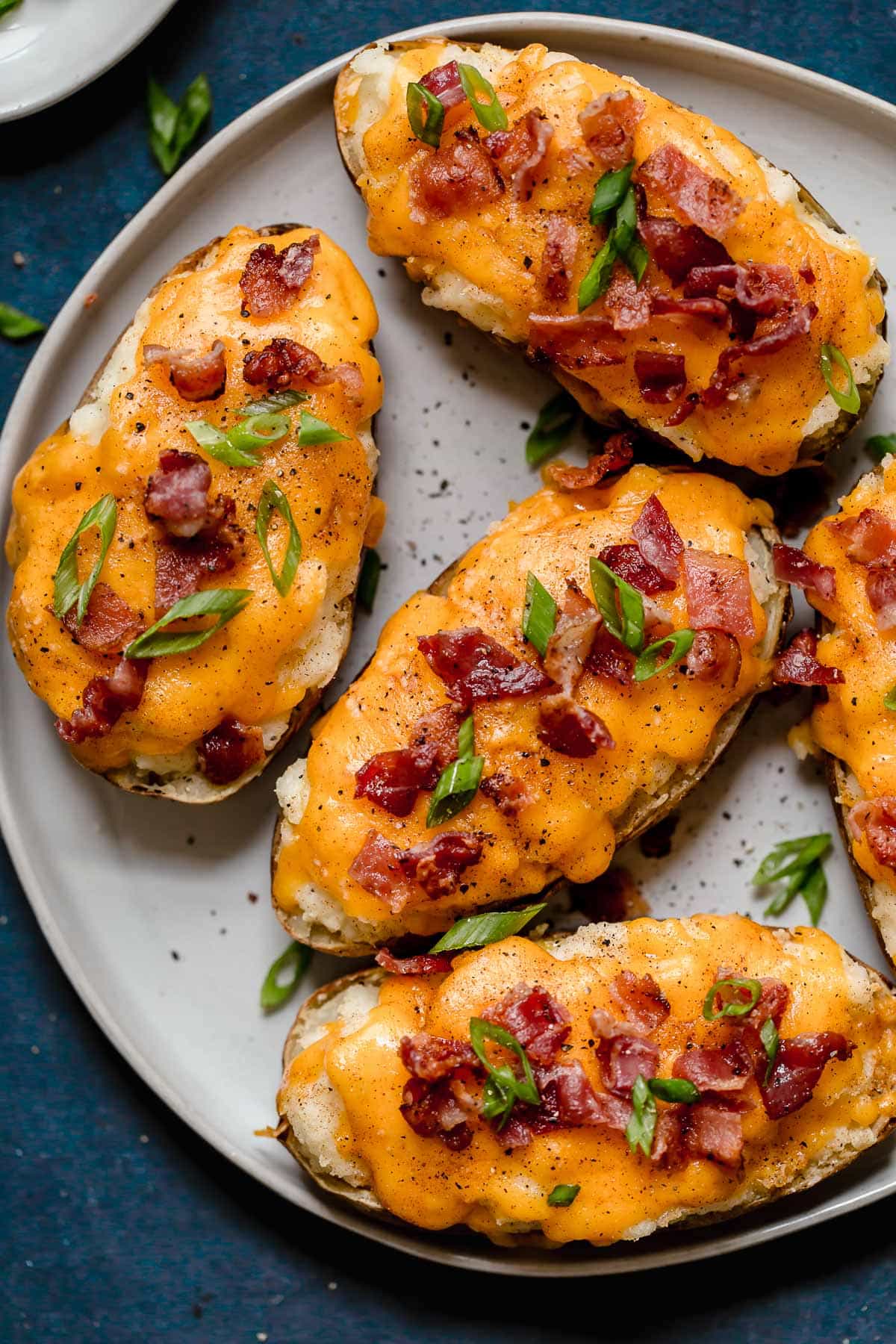 Overhead image of four Twice Baked Potatoes on a white serving plate. Potato skins are filled with mashed potatoes, topped with melted cheddar cheese and garnished with bacon and green onions.