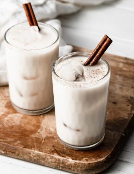 Horchata in two glass cups with ice cubes and a cinnamon stick. Cups are set over a wooden cutting board.