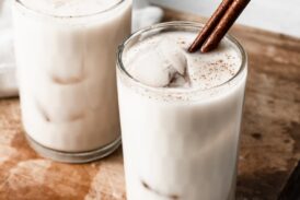 Horchata in two glass cups with ice cubes and a cinnamon stick. Cups are set over a wooden cutting board.