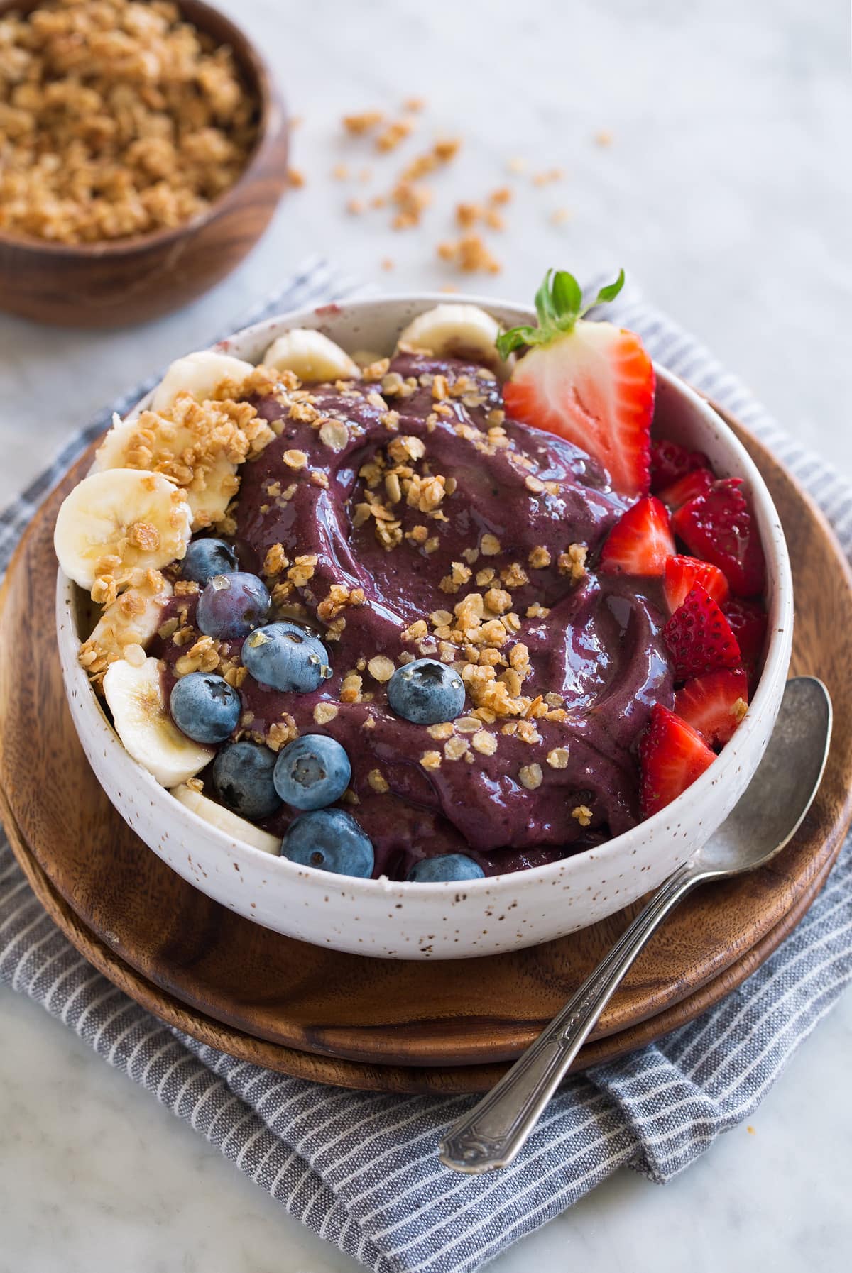 Thick acai bowl topped with strawberries, blueberries, bananas, granola and honey. Bowl is set over a wooden plate and a grey striped napkin.