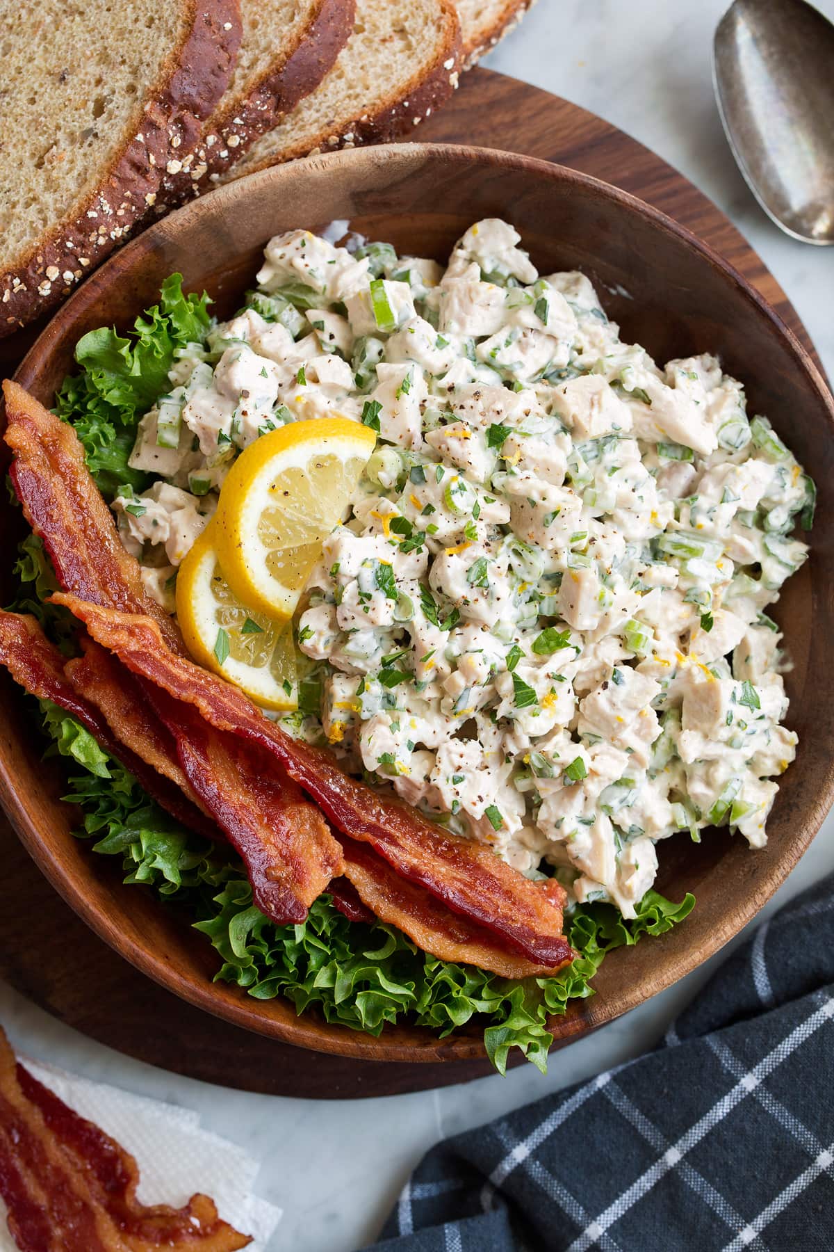 Chicken salad in a wooden bowl with a side of bacon and lettuce. It is garnished with lemon slices and there are slices of bread to the side of the bowl.