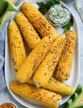 Eight grilled corn on the cob on a white oval platter with flavored butters on the side.