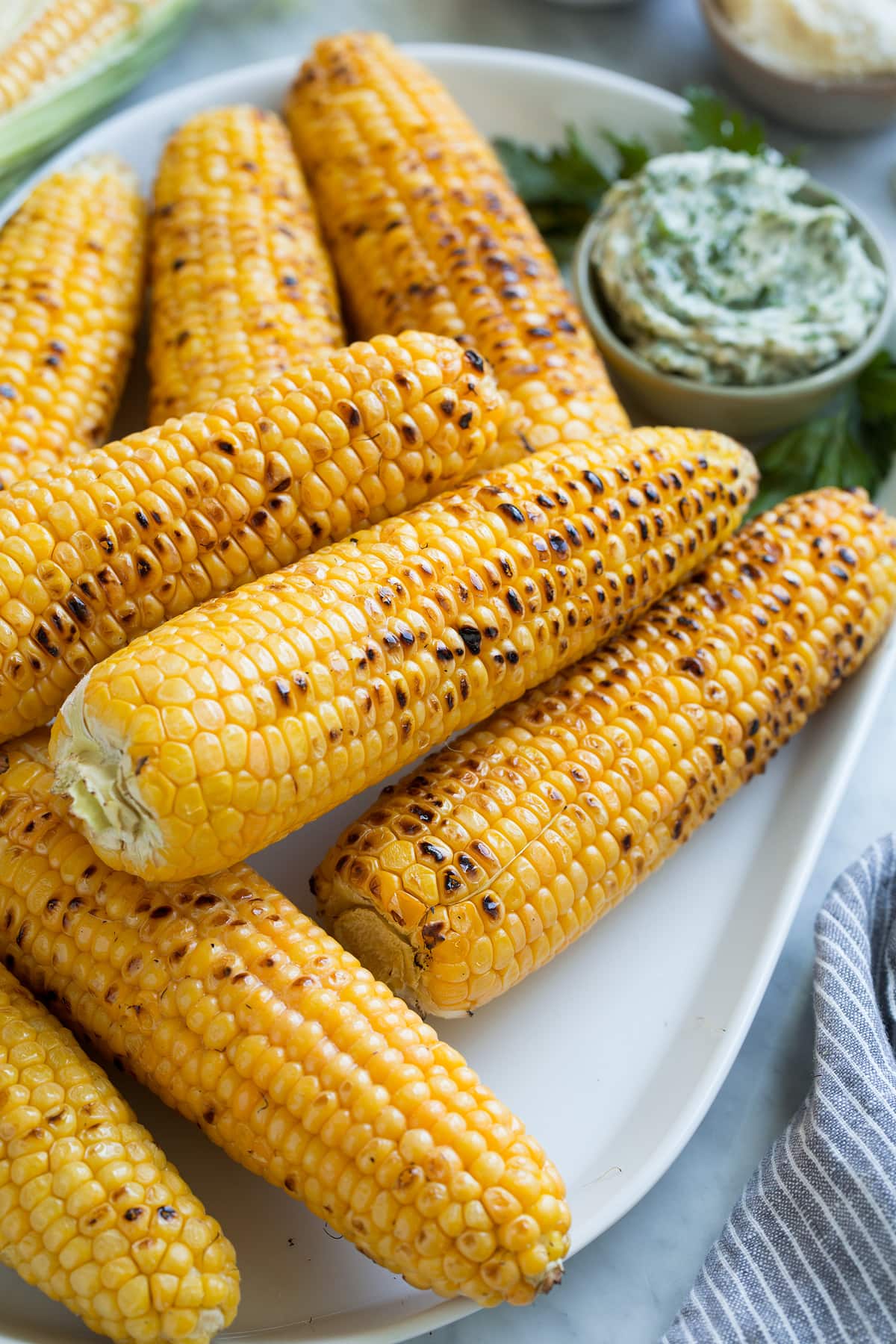 Close up image of stack of grilled corn on the cob on a white oval serving platter set over a marble surface.