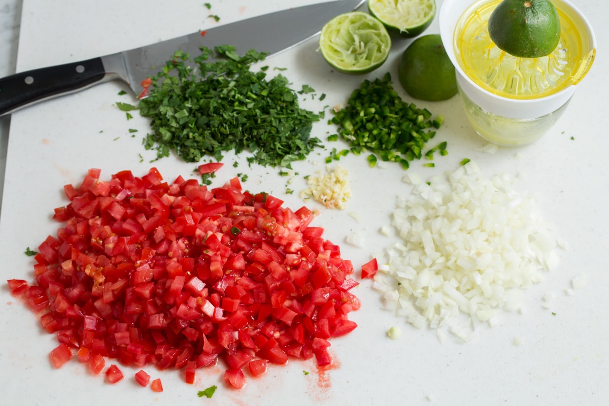 Finely chopped tomatoes, onion, cilantro, jalapeno pepper and garlic on a cutting board with a knife in the background and a juicer.