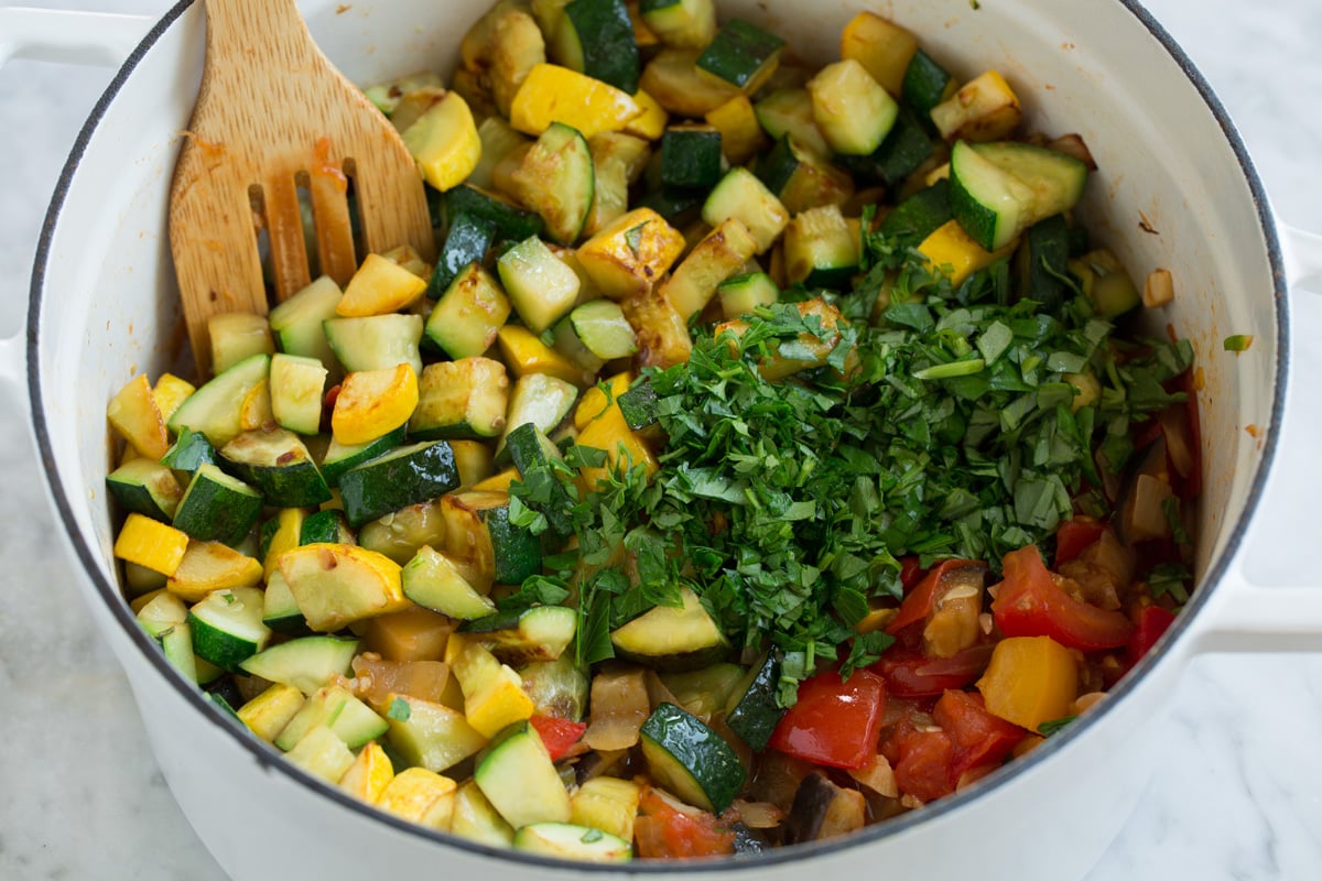 Adding squash and more fresh herbs to ratatouille mixture in pot.