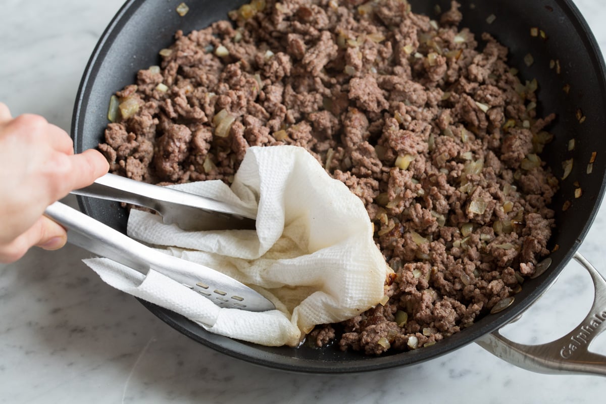 Draining fat from ground beef in skillet using a paper towel and tongs.