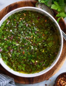 Overhead close up image of chimichurri in a bowl set over a wooden plate on a marble surface.