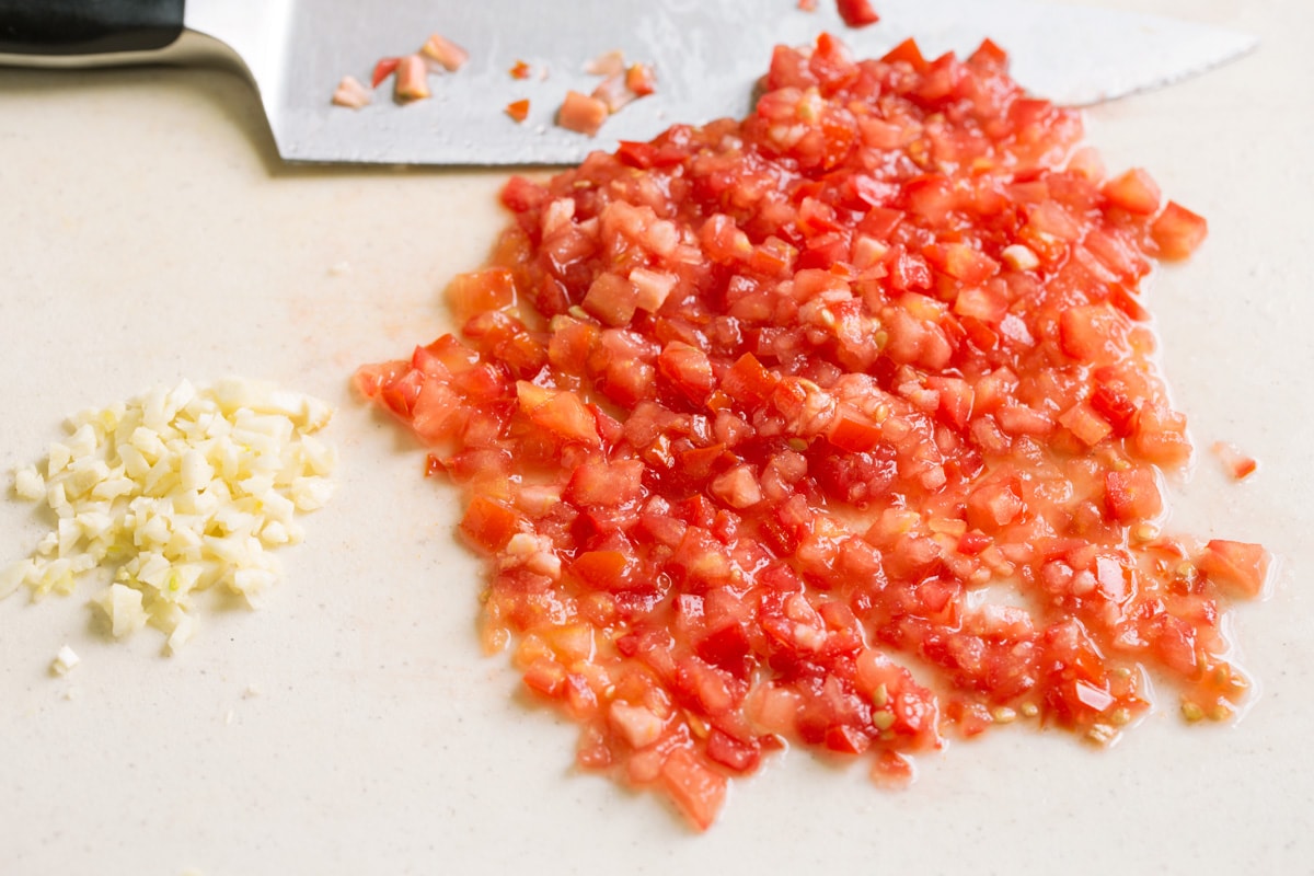 Finely chopped tomatoes and garlic on a cutting board with a knife on the side.