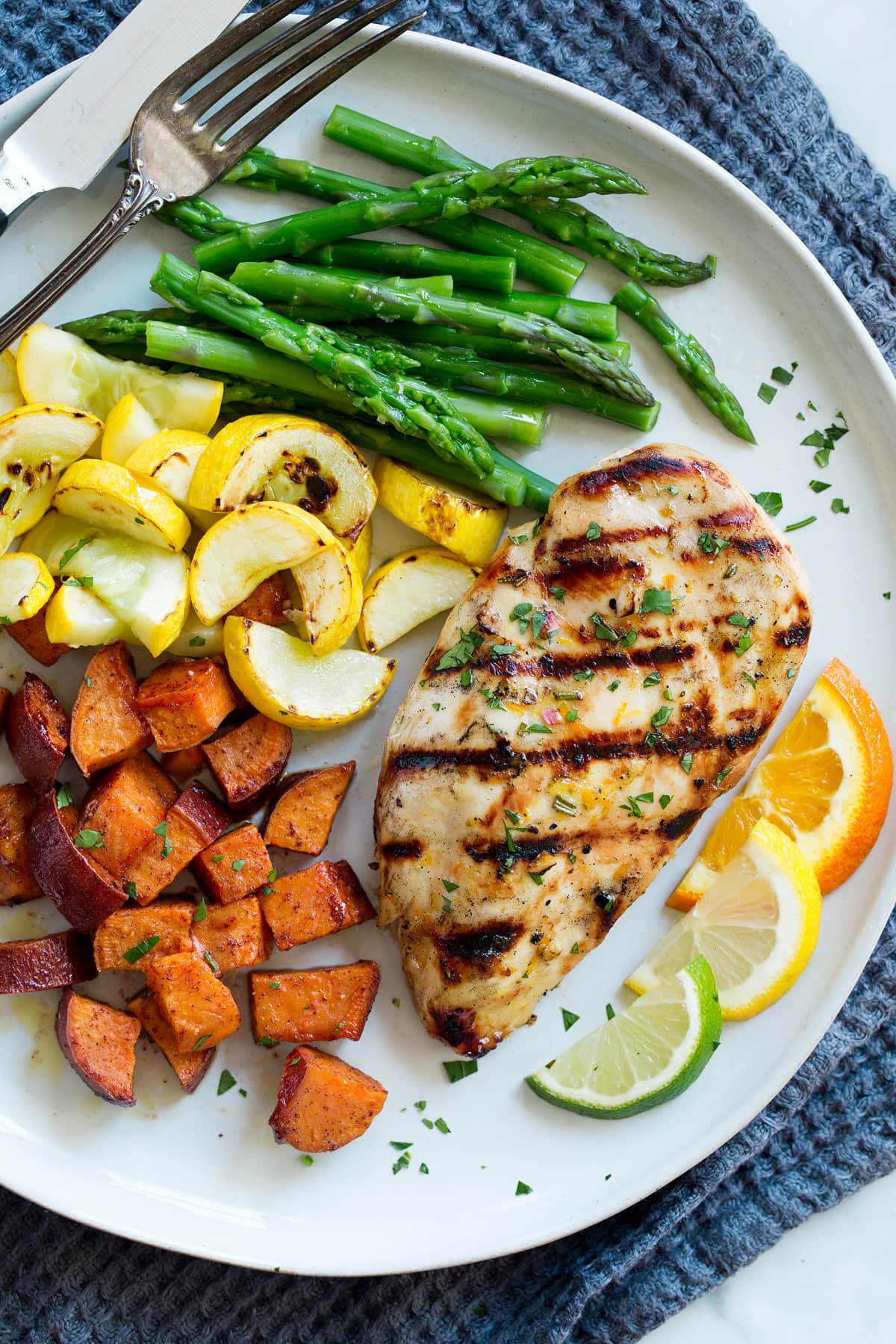 Marinated chicken on a serving plate with a side of asparagus, yellow squash, sweet potatoes and citrus slices.