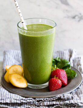 Green smoothie in a tall glass with strawberries, spinach and bananas to the side.