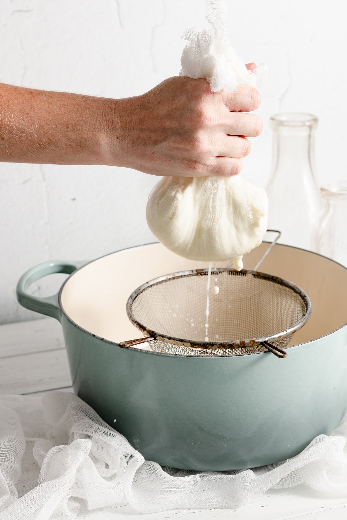 Hand holding cheesecloth full of ricotta cheese that is draining of liquid into a pot.