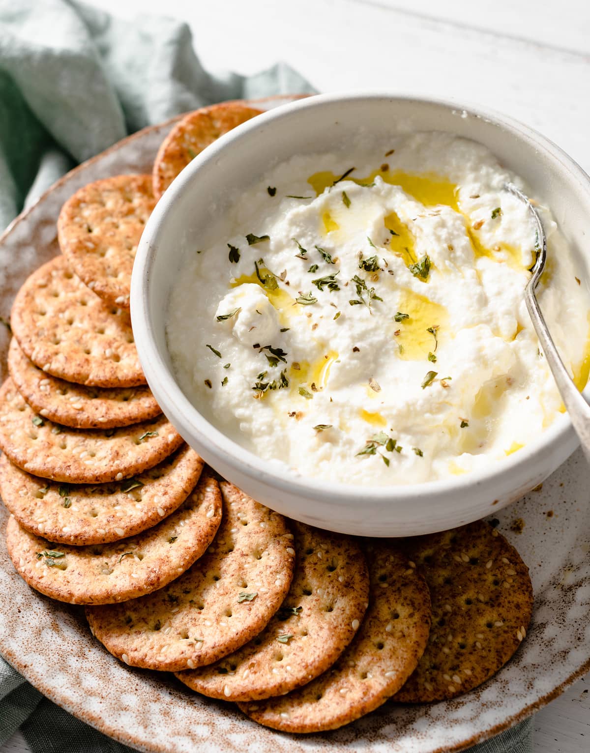Homemade ricotta in a serving bowl drizzled with olive oil, sprinkles with dried herbs and served with a side of crackers.