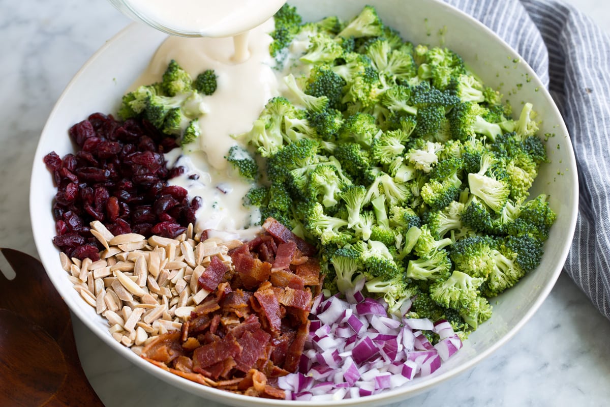 Pouring dressing over broccoli salad ingredients in a large white bowl.