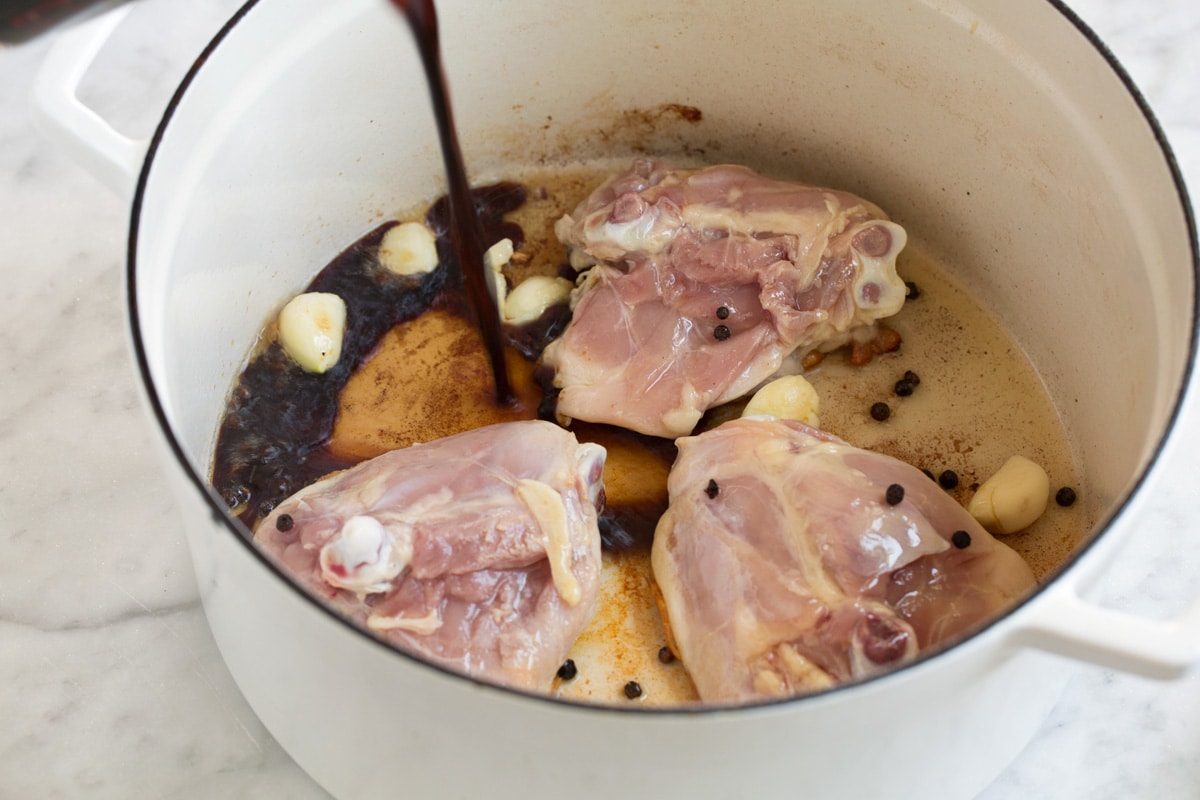 Pouring soy sauce and vinegar mixture into pot with chicken thighs, garlic and peppercorn.
