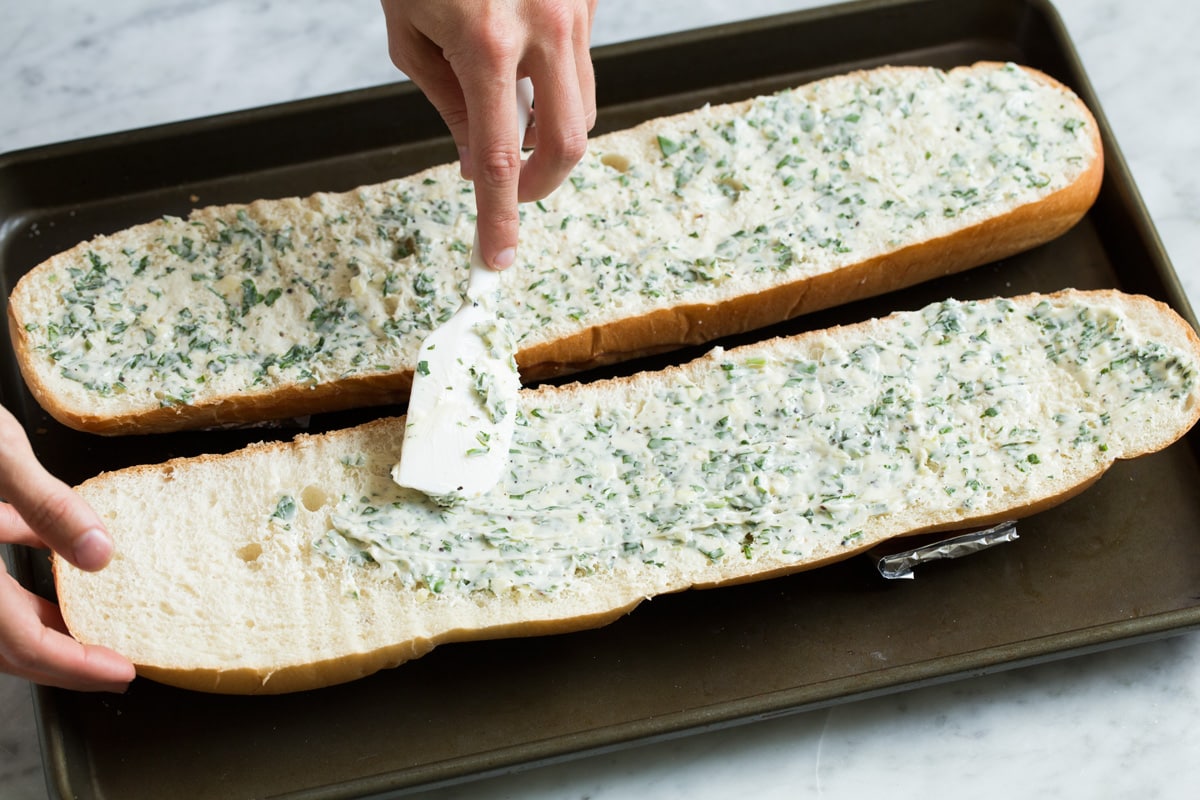 Spreading garlic butter over french bread halves on a baking sheet.