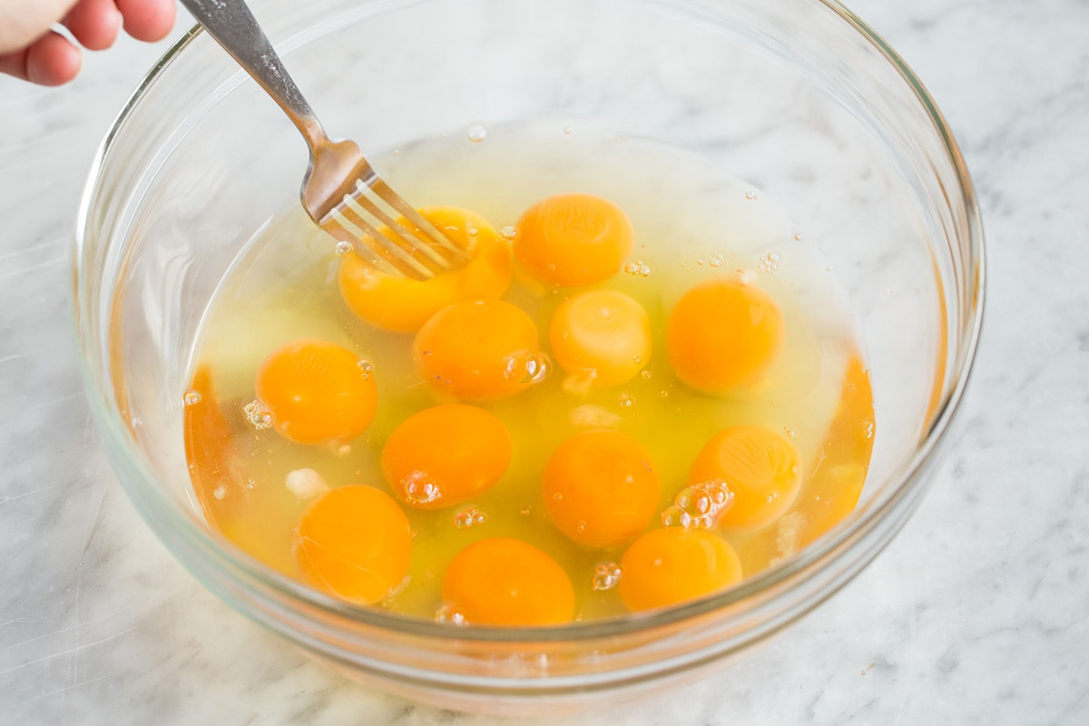12 eggs in a glass bowl, hand shown starting to mix with a fork.