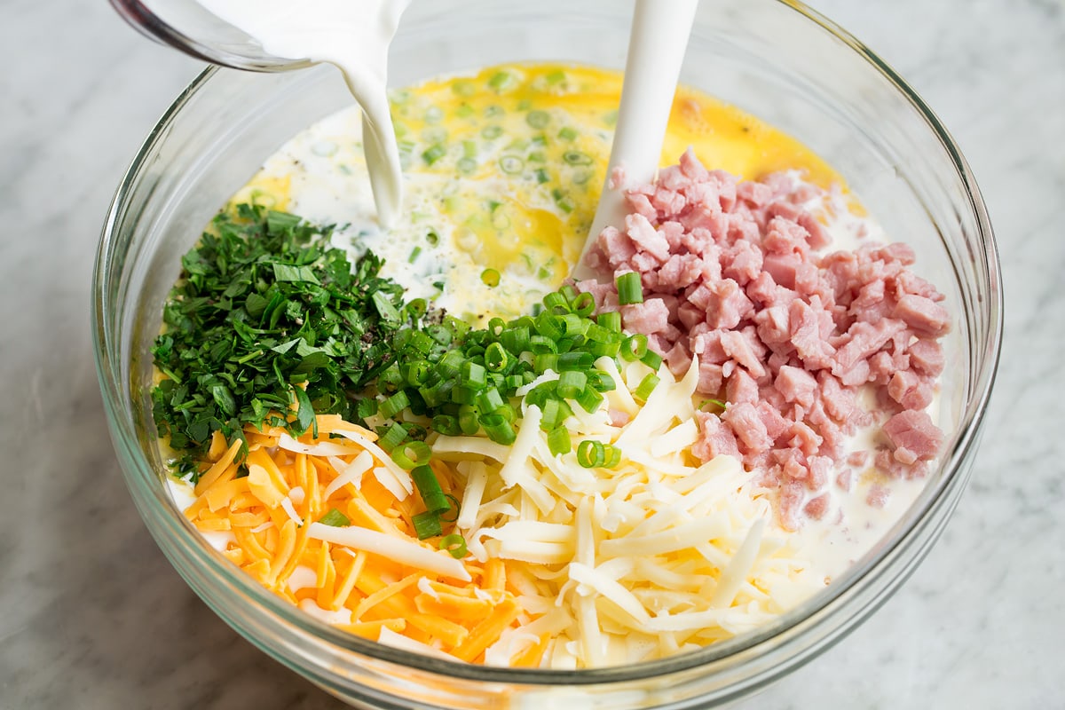 Pouring half and half into glass bowl filled with eggs, ham, shredded cheese, green onions and parsley.
