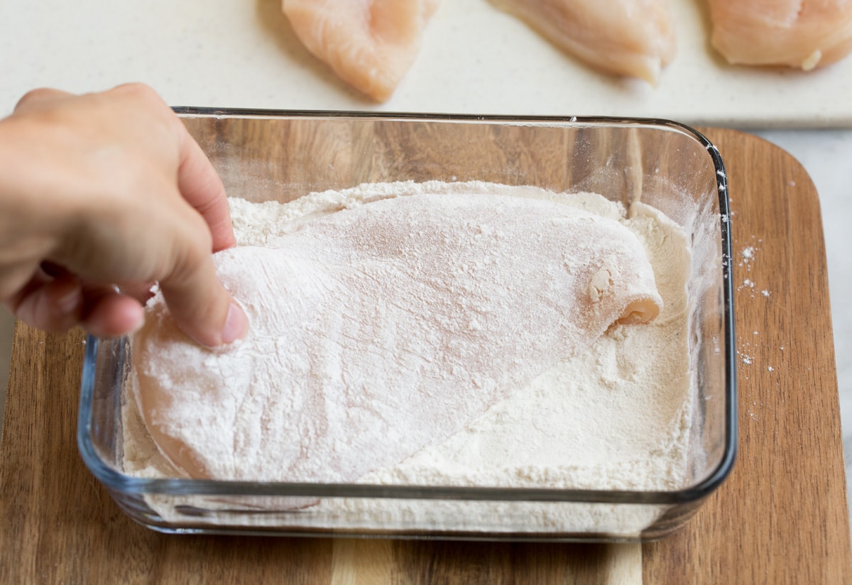 Dredging a chicken cutlet in flour in a glass dish.