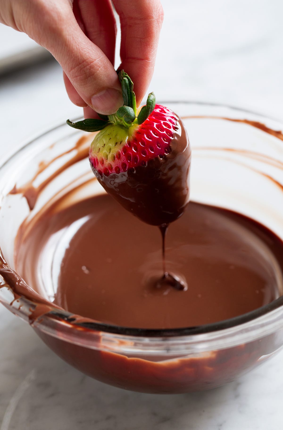 Hand holding strawberry by leaves and dipping strawberry in melted chocolate.