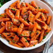 Photo: Roasted carrots in a white serving bowl. Carrots are garnished with parsley with a bunch in the background. Bowl is resting on a wooden platter and a grey cloth.