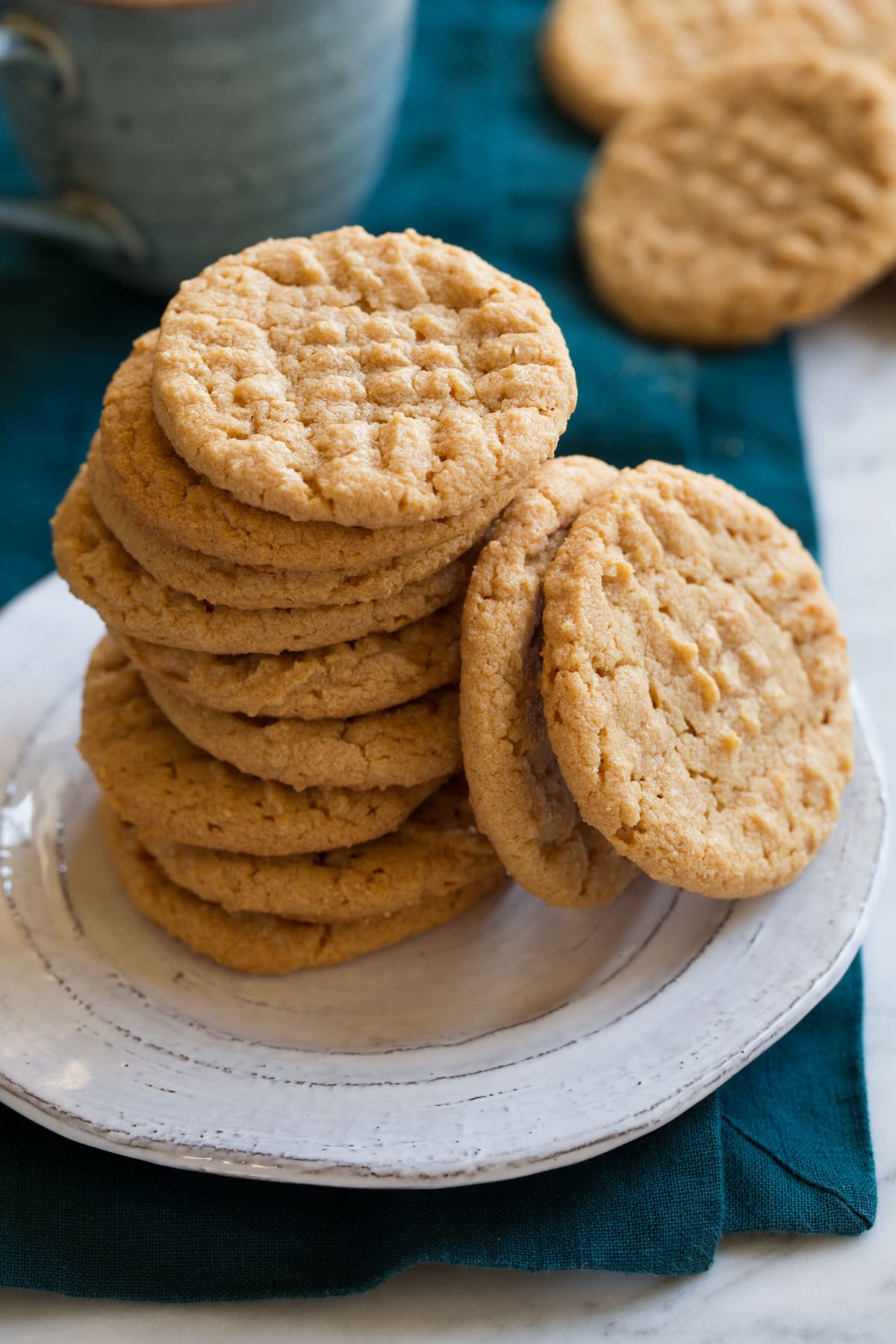 Three ingredient peanut butter cookies stacked on a white plate with a pile of cookies and mug in the background.