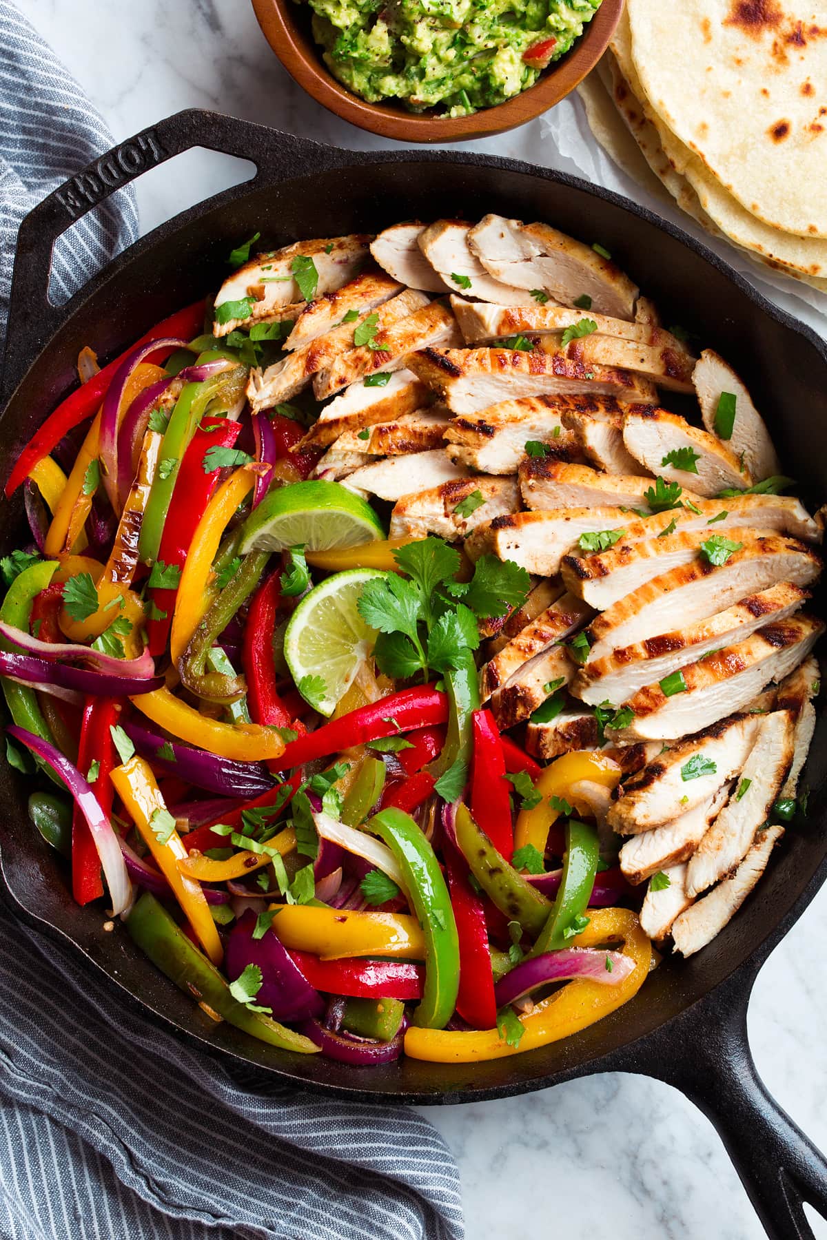 Chicken Fajitas mixture in a cast iron skillet. Showing sliced chicken breasts, bell peppers and onions. Tortillas and guacamole are served on the side.